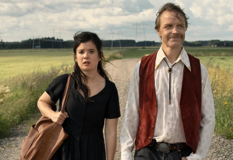 Kuvassa ovat Aksa Korttila mustassa mekossa ja olkalaukku vasemmalla  olallaan ja Jani Volanen valkoisessa paidassa ja violetissa liivissä. Kuva on puolivartalokuva. He kävelevät hiekkatietä pitkin ja ympärillä on kellertävää viljapeltoa. 