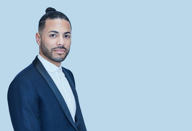 Kuvassa on Samu Moussa puolivartalokuvassa kuvan vasemmassa laidassa. Hänellä on pään päällä pieni nuttura ja päällään tumman sininen takki ja valkoinen paita. Tausta on vaalean sininen.