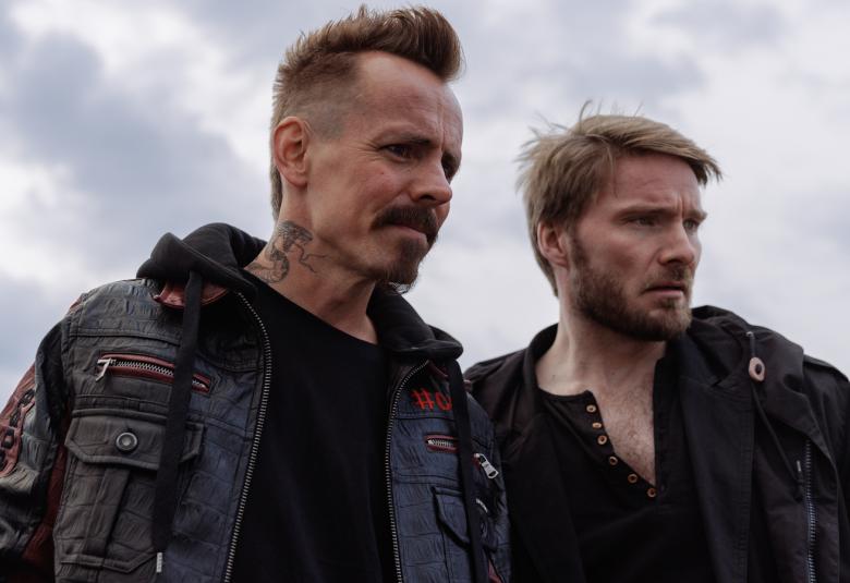 Kuvassa on vasemmalla Jasper Pääkkönen ja oikealla Leo Sjöman.  Heillä on tummat takit päällään ja he ovat puolivartalokuvassa ja katsovat alaspäin. Taustalla on harmaita pilviä.