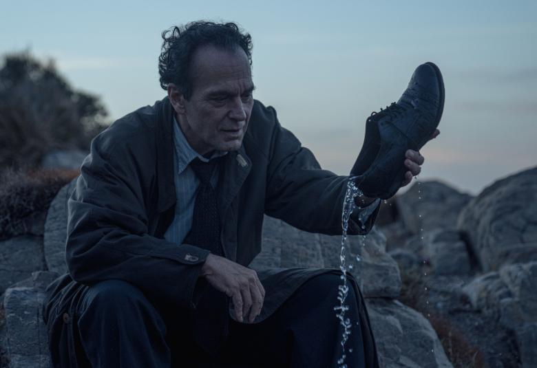 Kuvassa on mies kyykyssä kaaktamassa kengästään vettä. Taustalla näkyy pensas. Kuvan sävy oli tummahko ja kuvaa hämärää iltaa.