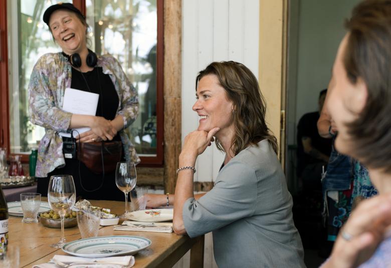 Kuvassa on Pirjo Lonka keskellä istumassa katetun päivällispöydän ääressä. Takana seisoo Ulla Heikkilä ja edellä on nainen selin epätarkasti kuvattuna.