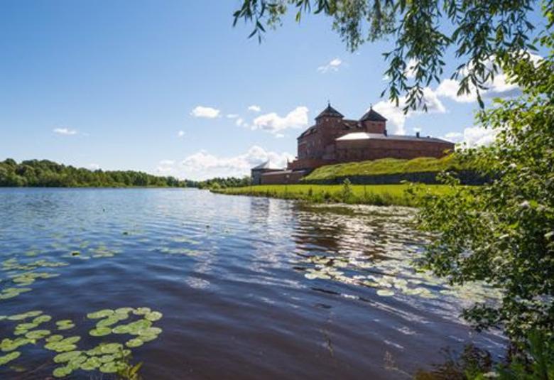 Kuvassa on Hämeen linna oikealla veden ääressä. Vedessä on ulpukan lehtiä. Vasemmalla näkyy lehtipuumetsää. Taivas on sininen ja siinä on pari pientä poutapilveä. 