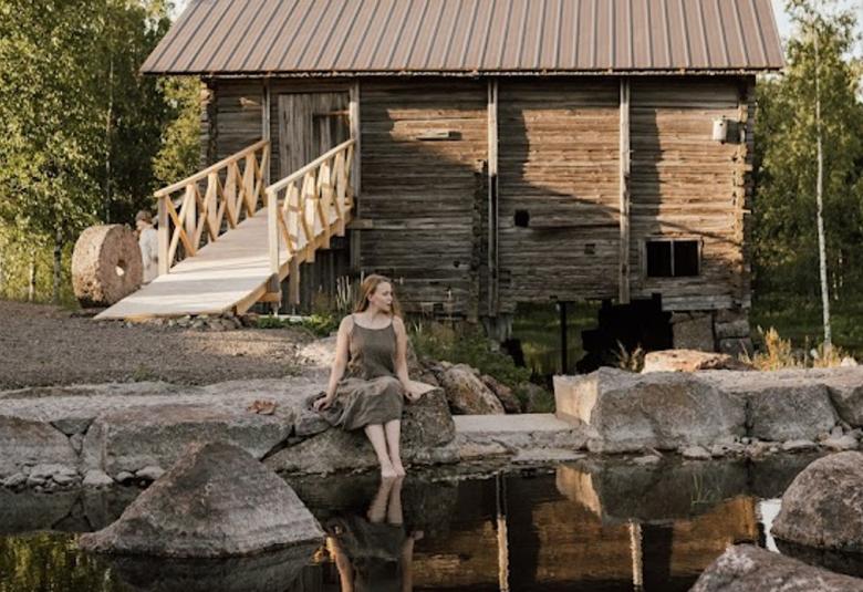 Kuvassa on takana harmaa hirsinen myllyrakennus. Sinne johtaa vetosilta ja edessä on laakeaa harmaata kiveä. Edessä on vettä ja harmaan kivipaaden päällä istuu nainen ruskeassa hameessa. Toisella puolella on myös kivipaasirakennelma.