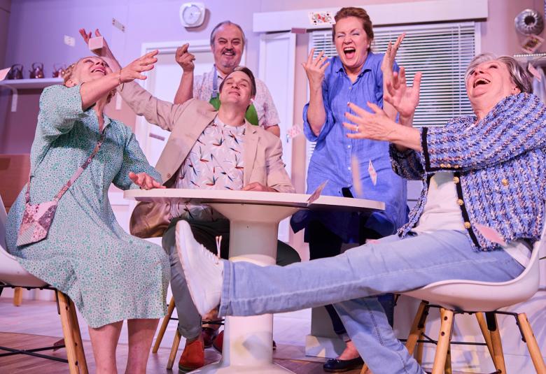 Kuvassa ovat kaikki näyttelijät valkoisen pöydän ääressä ja he nauravat sekä ojentelvat käsiään. Kolme istuu ja kaksi seisoo pöydän takana.