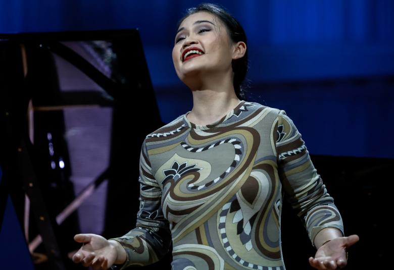 Kuvassa on Jingjing Xu laulamassa. Hän on puolivartalokuvassa ja hänellä on vihreä kuviollinen pitkä hihainen mekko päällään.