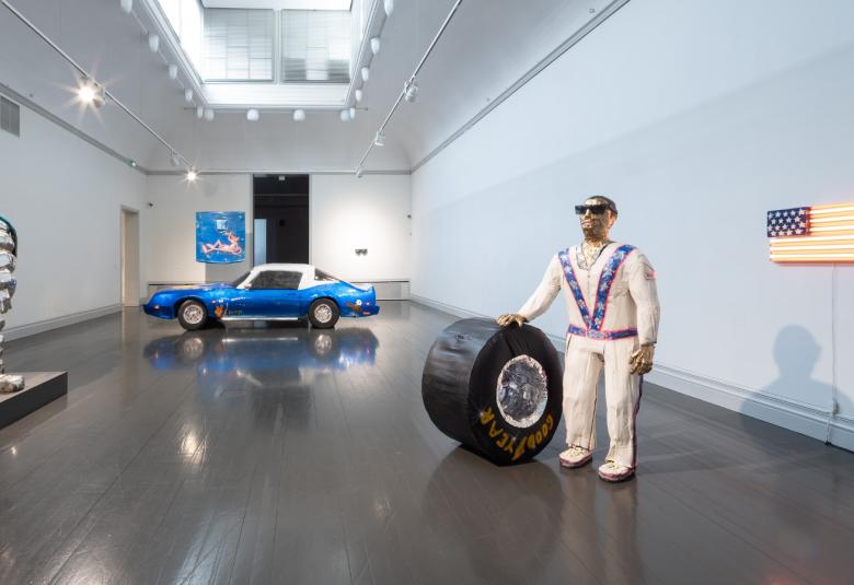  Kuvassa on tilassa, jossa on kiiltävä harmaa lattia ja valkoiset seinät ja katto.  Vasemmalla on valkoisen harmaa avaruuspukumainen teos. Oikealla on valkoisessa toppapuvussa aurinkolaseissa mies ja hän pitää oikealla kädellään leveää rengasta. Miehen takana on USA:n lippu. Takana on sininen, valkokattoinen auto.