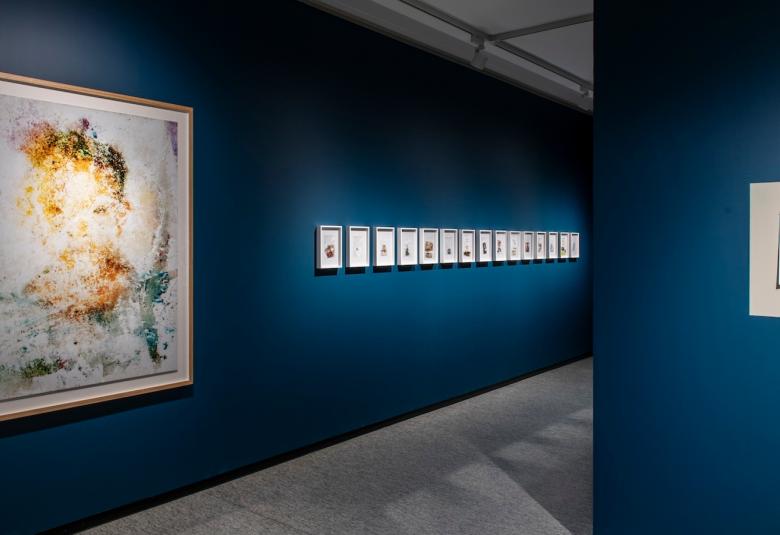 Kuvassa on taidenäyttelyn käytävää ja siinä on sinisellä seinällä vasemmalla iso teos.  Siinä näkyy kellertävät kasvot.  Oikealla on rivi pieniä teoksia.