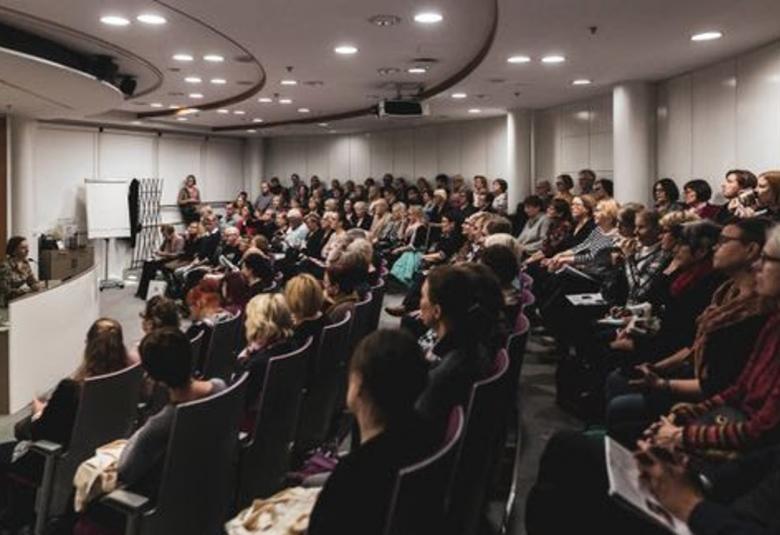 Kuvassa on auditorio, jossa on vasemmalla korkeammalla tasolla olevan pöydän ääressä nainen puheenjohtajana.  Hänen edessään on puolikaaressa yleisöä.