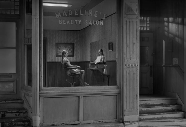 Kuvassa on ulkoapäin harmaan tummaa rakennuksen seinää ja ukkunasta näkyy kaksi naista istumassa pöydän molemmin puolin. Vasemman puoleisella on käsi pöydällä.  Ikkunan päällä lukee Madeleines Beaauty Salon