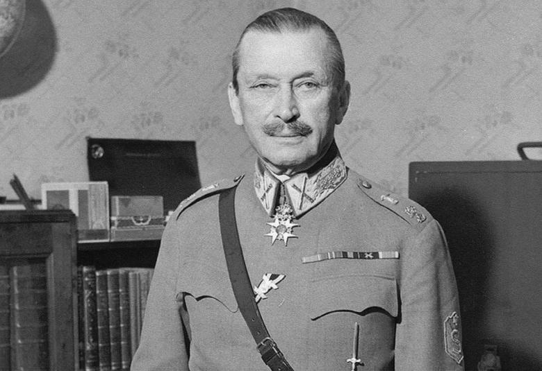 Kuvassa on Mannerheim puolivartalokuvassa ja hänellä on sotilaspuku päällään.  Taustalla on toimistohyllyä. Kuva on musta-valkoinen.