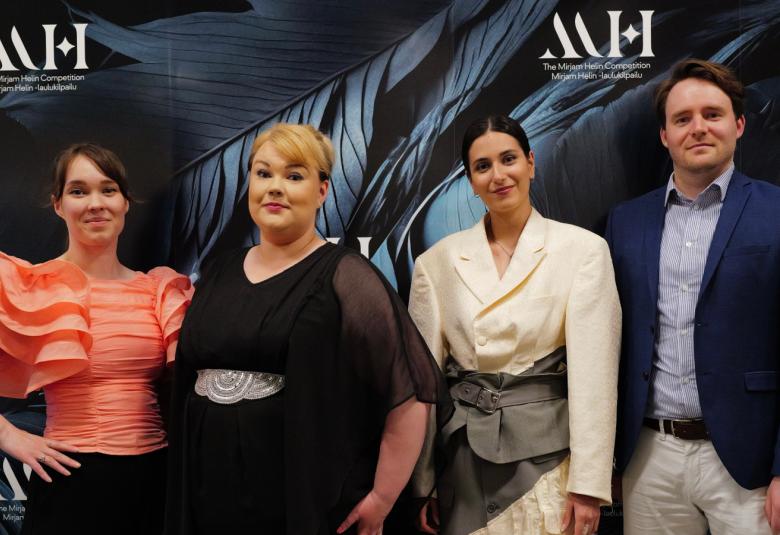 Kuvassa ovat vasemmalta oranssissa, röyhelöisessä puserossa ja mustassa hameessa, Heta Sammalisto mustassa mekossa, Iris Candelaria valkoisessa mekossa ja Johannes Pessi tumman sinisessä takissa ja vaaleissa housuissa.  Tausta on tumma ja siinä on ylhäällä MH-kirjaimet kolmesti.