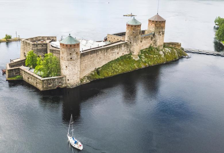 Kuvassa on ilmasta kuvattuna Olavinlinna veden keskellä ja siinä näkyy kolme tornia ja kivimuuria. Ympärillä on vettä ja reunoilla näkyy hiukan vihreää puustoa. 