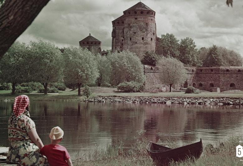 Kuvassa on Olavinlinna takana sen edessä hopeapajuja. Edessä istuu  selin nainen mekko päällään ja huivi päässään katsomassa linnaa. Hänen oikealla puolellaan on poika vaalea pipo päässään. Linnan ja henkilöiden välissä on vettä. Oikeassa nurkassa on luku 550. Kuva on musta-valkoinen.