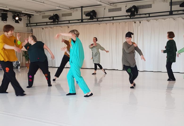 Kuvassa ovat tanssijat erilaisissa asennoissa ja vauhdikkaissa liikeessä. Edessä on nainen turkoosissa koko vartalopuvussa. Muilla on mustia housuja ja eri värisiä puseroita.