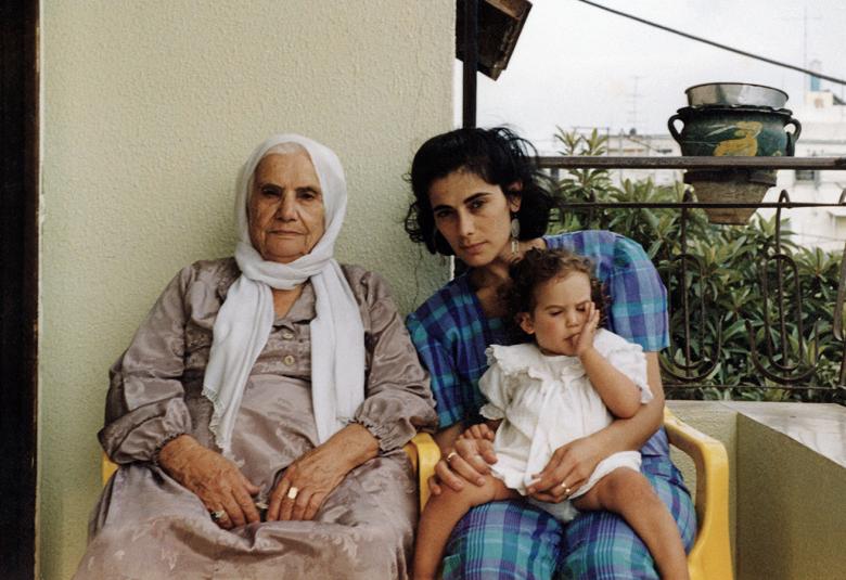 Kuvassa on parvekkeellä istumassa vanha nainen valkoisessa huivissa ja vaalean ruskeassa mekossa. Hänen vieressään oikealla istuu nuori nainen siniruutuisessa mekossa ja hänen sylissään on pikku tyttö valkoisessa mekossa. Takana on osittain vaaleaa seinämää ja oikealla vihreää oksistoa.