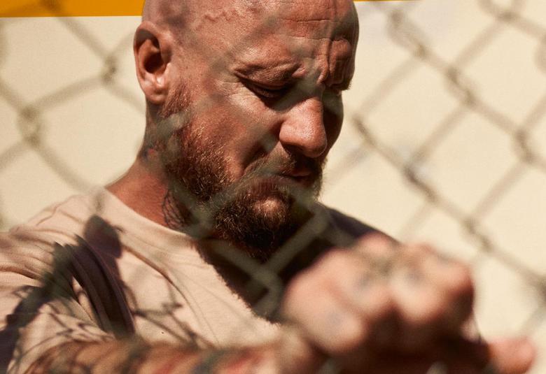 Kuvassa on ruskettuneen miehen kasvot ja hän ojentaa nyrkkiään. Hänellä on vaalean ruskea paita. Edessä näkyy metalliverkkoa. Kuva on osa kirjan kannesta ja se on kasvokuva.