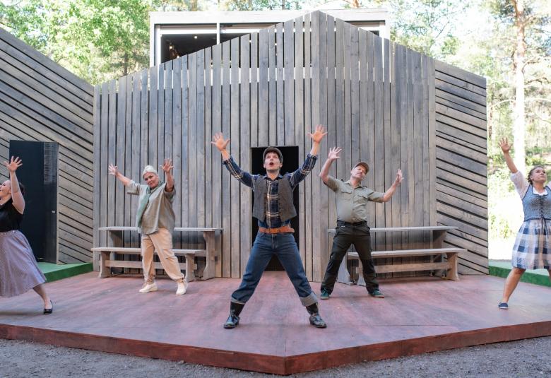 Kuvassa on kolmen harmaan seinän vinotetulla näyttämöllä tanssiesitys, jossa kaikilla näyttelijöillä on kaikilla kädet ylhäällä. Reunoilla on kaksi naisnäyttelijää hameet päällään ja kolme miestä keskellä eri värisissä vaatteissa.