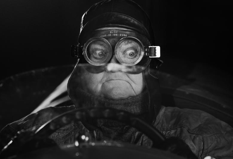 Kuvassa on mies vanhanaikaisessa moottoripyörävarustuksessa nahkaisessa kypärässä. Tausta on musta.