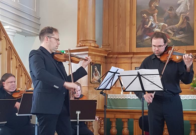 Kuvassa ovat vasemmalla Jukka Merjanen ja Petteri Iivonen soittamassa viulua Haminan Johanneksen kirkon alttarilla. Taustalla näkyy osa alttaritaulusta.