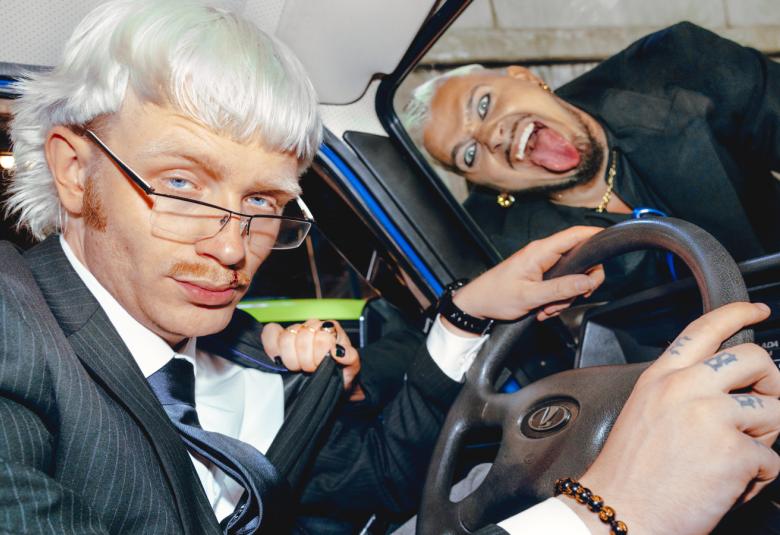 Kuvassa on vaaleatukkainen Joost tummassa pikkutakissa kädet autonratissa. Kuva on otettu auton sisältä.  Käärijä on vinottain auton tuulilasin päällä tummassa nahkatakissa ja näyttää kieltään.