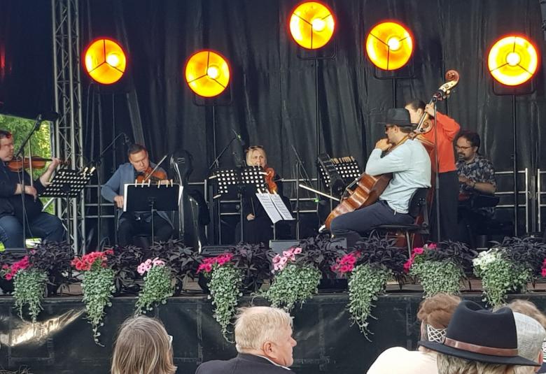 Kuvassa ovat soittajat lavalla ja lavan reunassa punaisia kukkia ja vihreitä roikkuvia kasveja. Etualalla on alhaalla yleisöä. Lavalla ovat soittamassa vasemmalta Ville ja Matti Koponen, Eriikka Nylund ja edessä Tuomas Ylinen vaaleassa paidassa ja lierihatussa. Hänen takanaan punaisessa pusakassa on Maria Krykov ja takana Tuomas Turriago.  Tausta on musta ja siinä on keltaisia pyöreitä valoja.