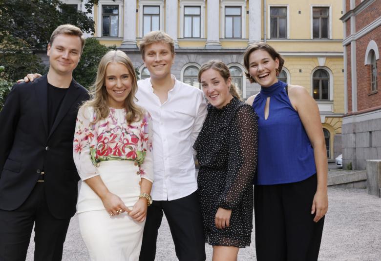 Kuvassa on ryhmä nuoria ja vasemmalla on mies mustissa vatteissa. Vieressä on nainen punakukallisessa paidassa valkoisessa puolihameessa. Kesekellä on mies valkosiessa paidassa ja vieressä nuori nainen tumman sinisessa mekossa ja toinen sinisessä puserossa. Taustalla on kelta-valkoinen rakenus klassisine pylväineen.