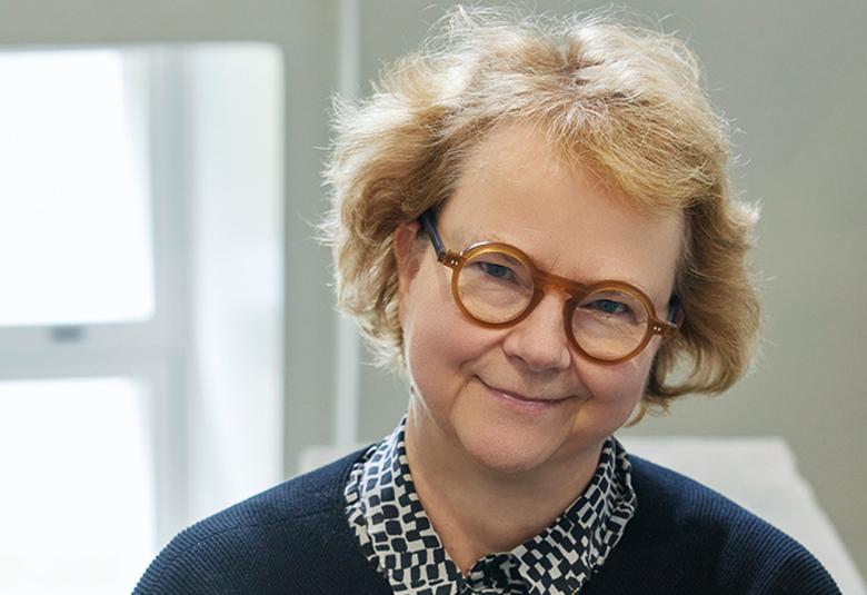 Kuvassa on Laura Kolbe kasvokuvassa ja hänellä on vaaleat hiukset ja ruskeat silmälasit .  Päällään hänellä on sininen villatakki sekä sini-valkoruudullinen paita. 