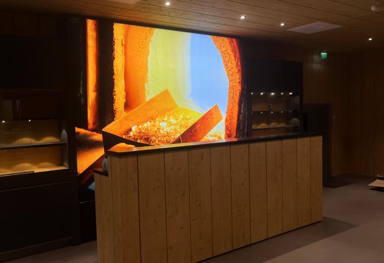 Kuvassa on ruskean kaapin päällä ahjon leveä suu, josta näkyy tulta ja edessä lasimassaa. Tausta on mustaa. 