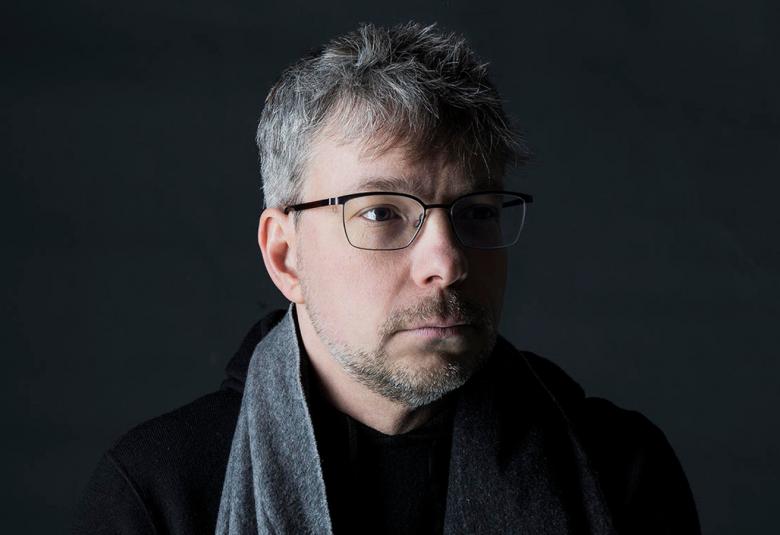 Kuvassa on Olari Elts kasvokuvassa ja hänellä on katse jonkin verran oikealle. Hänellä on silmlasit ja musta paita päällään sekä oikean olkapään yli tuleva harmaa kaulahuivi. Tausta on musta.