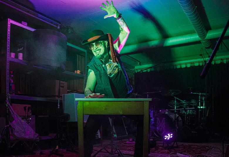 Kuvassa on mies rummuttamassa pöytää ja hänellä on vasen käsi ylhäällä.  Päässään hänellä on stetson ja päällään musta liivi ja t-paita. Taustalla on tummaa hyllyä.  