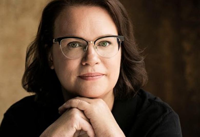 Kuvassa on Sirpa Kyyrönen, jolla on olkapäille ulottuvat pitkät hiukset ja silmälasit.  Hän pitää käsiään leuan alla ja kuva on puolivartalokuva. Tausta on puoliksi ruskean sävyinen ja puoliksi musta.