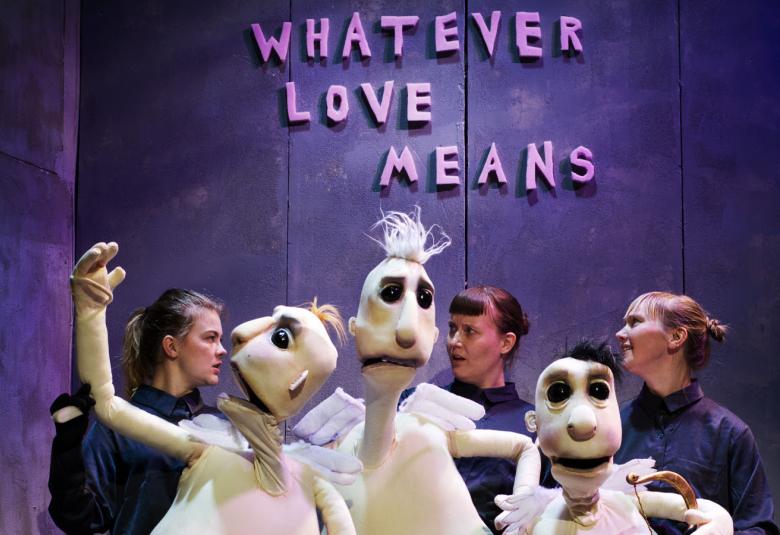 Kuvassa on edessä valkoisia isoja mieshahmoisia pehmonukkeja.  Takana on kolme naisnäyttelijää, joilla on tummat vaatteet. Taustalla on tumman violetilla seinällä vaalean violetilla Whatever love means.