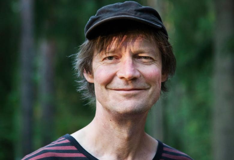 Kuvassa on Juha Hurme puolivartalokuvassa ja hänellä on musta lippalakki sekä raitapaita päällään. Taustalla on metsää.