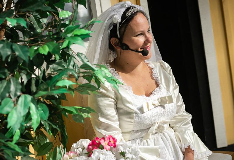 Kuvassa on vasemmalla vihreälehtinen kasvi ja sen vieressä istuu nainen valkoisessa morsiuspuvussa huntuineen.  