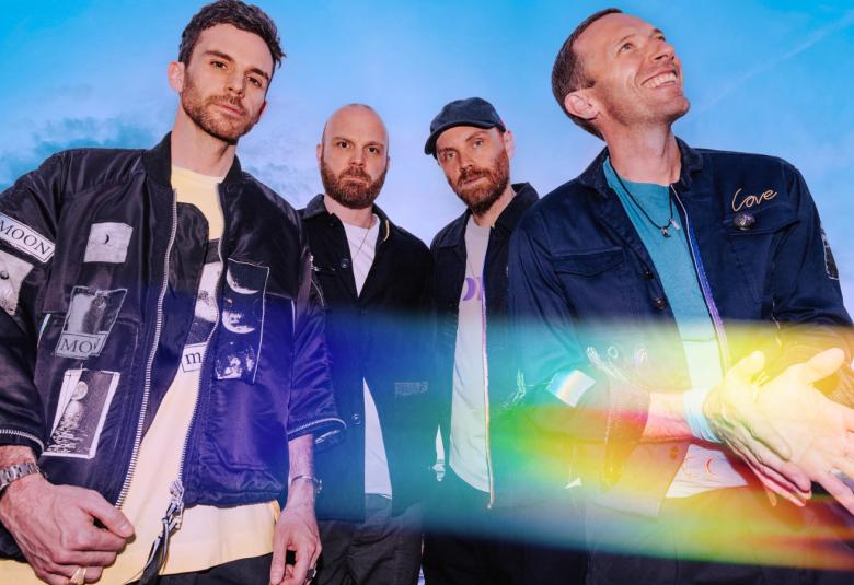 Kuvassa on neljä Coldplayn jäsentä ja heillä on tummat pusakat. Oikealta alhaalta tulee hieman kelta-sinistä valoa. 