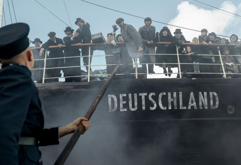 Kuvassa on laiva, jonka kyljessä lukee Deutschland.  Laivan kannella kaiteen takana on ihmisiä tummissa vaatteissa. Vasemmalla on mies selin tummassa poliisin asussa koppalakissa pyssy kädessään.