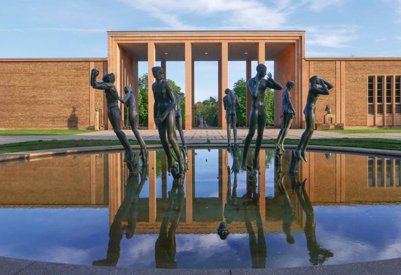 Kuvassa on pyöreän vesialtaan takana on ruskea, molemmin puolin laatikkomaiset rakennukset, joiden keskella laatikkomainen pylväikkö. Taivas on sininen ja se heijastuu altaan vedenpitaan samoin kuin rakennus pylväikköineen. Altaassa on ympyrässä ihmispatsaita.