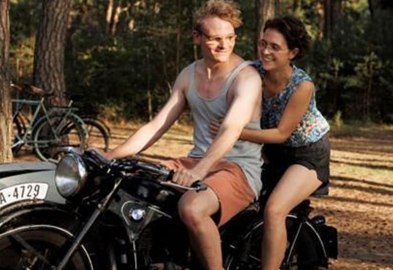 Kuvassa ovat moottoripyörän päällä nuori mies ja hänen takanaan on nuori nainen istumassa pyörän takapenkillä.  Taustalla on metsää.