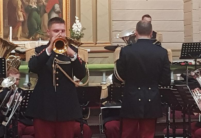 Kuvassa on flyygelitorven soittaja etualalla mustassa kultanappisessa takissa ja taustalla on selin kapellimestari sekä taustalla soittajia. Takana näkyy osa Mäntyharjun kirkon alttaritalua.