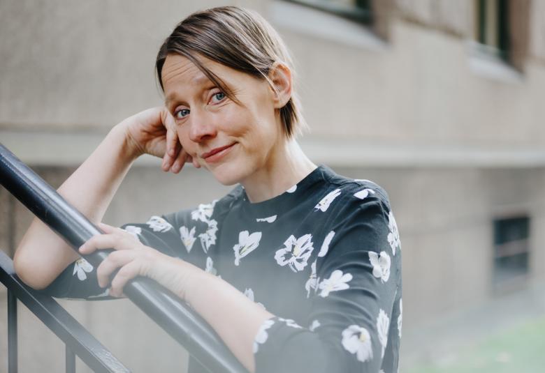 Kuvassa on Ingeborg Arvola, josta on puolivartalokuva.  Hän nojaa ylöspäin menevään metallikaiteeseen oikealla  kyynerpäällään ja pitää vasenta kättään kaiteessa.  Hänellä on sininen valkokuvioinen mekko päällään. Taustalla näkyy vaaleaa matalaa taloa.