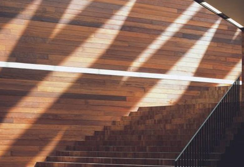 Kuvassa on ruskeaa lautaseinää ja portaikko. Keskellä seinää menee valkoinen lauta. Ylhäältä tulee valoa katon ja seinän rajassa olevista valoaukoista.