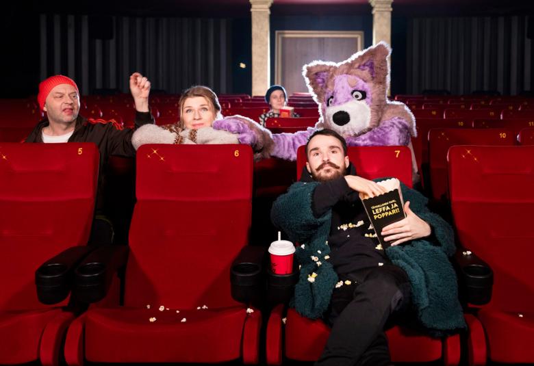 Kuvassa on punaisten istuimien katsomossa eturivissä yksi mies mustissa vaateissa ja hän on rennosti vieressään kahvimuki ja syö popcorneja. Hänen takanaan seuraavalla rivillä on vasemmalla mies tonttulakissa vasen käsi pystyssä ja hänen ieressään nainen nojaamassa tuolin yläosaan.  Hänen vieressään on vaalean violetin värinen koirapuvun pää ja siinä on valkea kuonon pää.