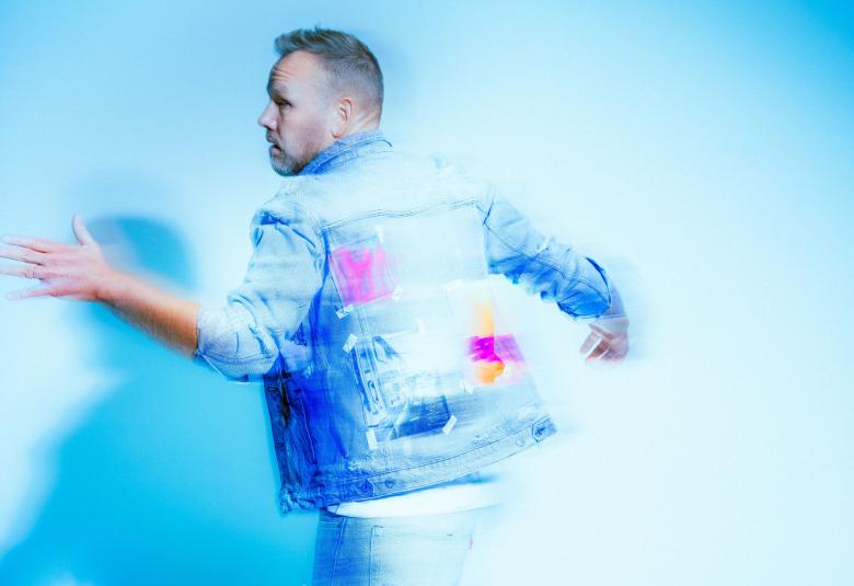 Kuvassa on tausta vaalean sininen ja Osmo Ikonen on vinottain oikealla vasen käsi ojennettuna eteenpäin. Hänellä on vaalean sininen farkkutakki päällään.