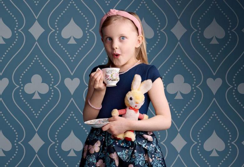 Kuvassa on pikkutyttö nalle kainalossa ja pieni kahvikuppi oikeassa kädessään. Hänellä on päällään mekko,jossa on musta yläosa ja kukallinen alaosa. Taustalla on sinistä tapettia, jossa on harmaita pataässiä ja hiitä on reunustettu samanmuotoisia koristeviivoituksia.