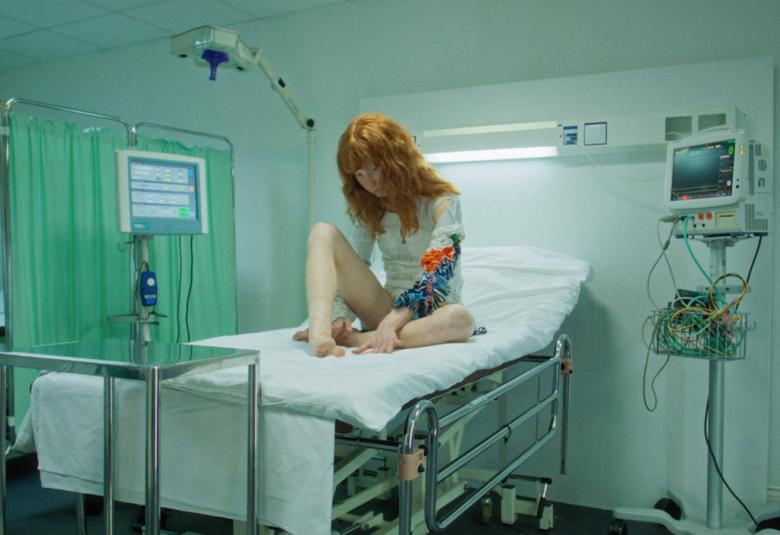 Kuvassa on sairaalasängyllä istumassa punatukkainen tyttö ja vasemmassa kädessään punainen ja sininen harmaan puseron hihan päällä kohokiviota. Vasemmalla on vaalean vihreää verhoa. 