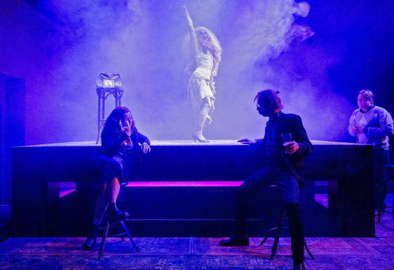 Kuvassa on sinisessä valossa näyttämöllä valkoisessa asussa nainen tanssimassa ja edessä katsojaan päin istumassa penkillä nainen vasemmalla ja oikealla mies. Esityslavan takana oikealla on mies.