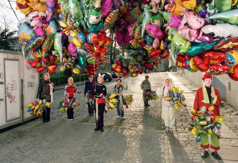 Kuvassa on nuoria pitelemässä runsasta määrää värikkäitä ilmapalloja vaalealla kadulla.