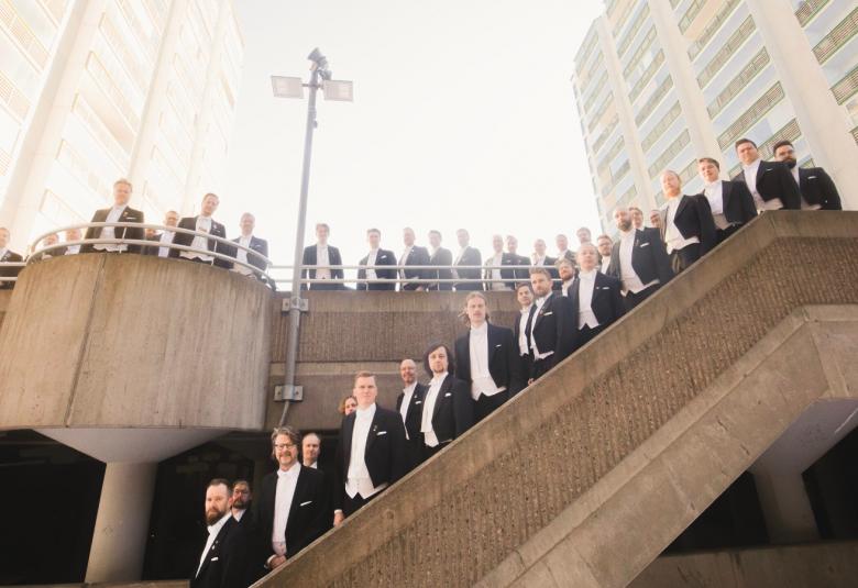 Kuvassa on Yloppilaskunnan Laulajat mustissa frakeissa ja valkoisissa paidoissa vasemmalta ylöspäin nousevissa portaissa ja ylhäällä tasanteella kaiteen takana. Kaiteet ovat harmaata betonia. Taustalla on vaaleata valoa.