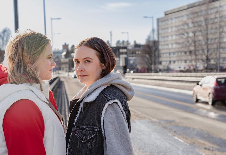 Kuvassa on vasemmalla kaksi tyttöä vastakkain ja heistä vasemmalla olevalla on valkoinen toppajakku ja punainen huppari. Oikealla olevalla on farkkuliivi ja harmaa huppari. Taustalla näkyy katua ja oikealla iso toimistorakennus.