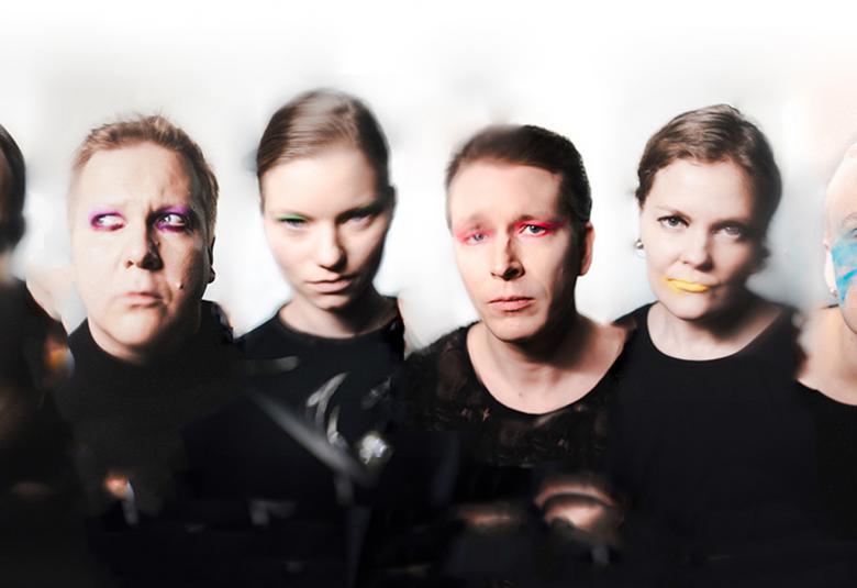 Kuvassa on kuusi näyttelijää puolivartalokuvassa ja heillä kaikilla on mustat puserot.  Oikealta toiseksi viimeisellä naisella on keltaiset huulet ja reunimmaisella miehellä turkoosia poskessaan.